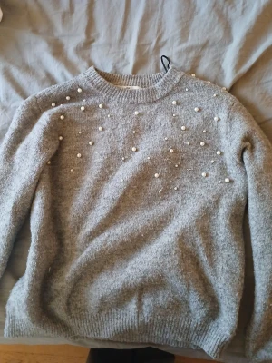 Grå stickad tröja med pärlor H&M - Snygg grå stickad tröja från H&M i storlek 146/152. Tröjan har rund hals och är dekorerad med pärlor över axlar och bröst för en extra cool look. Perfekt att matcha med jeans eller kjol. Långärmad och mjuk i materialet.