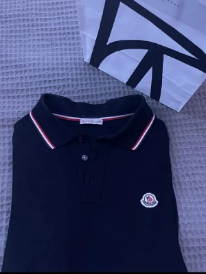 Svart Moncler polotopp med röd/vit kant - Säljer en svart polotopp från Moncler med klassisk krage och knappar framtill. Toppen har röda och vita ränder längs kragen och ärmsluten samt Moncler-logga broderad på bröstet. Materialet är bomull och modellen är kortärmad.