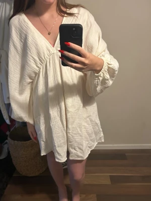 Vit sommarklänning - Säljer en luftig vit kortklänning med v-ringning och ballongärmar. Klänningen har en snygg öppen rygg med snörning och är gjord i ett mjukt bomullstyg med struktur. Perfekt för varma dagar och enkel att styla med accessoarer.