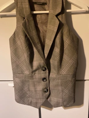 Grå rutig väst från Vero Moda - Snygg grå väst från Vero Moda i r sopassande rutig design. Västen har klassisk krage, tre knappar framtill och justerbart spänne i ryggen. Perfekt för att lyfta din outfit med en stilren touch.