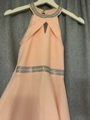 Ljusrosa långklänning med paljettdetaljer - Säljer en exakt likadan som jag inna la ut, denna är en mindre storlek bara. Denna är för 12 åringar. Men funkar för äldre. Också använd en gång. Säljer en elegant ljusrosa långklänning med glittrande paljettdetaljer vid halsringningen och midjan. Klänningen har ärmlös design, öppen rygg och ett snyggt nyckelhålsutskärning framtill. Materialet känns lätt och flowy, perfekt för speciella tillfällen.