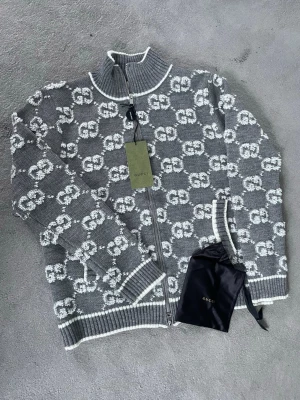 Grå stickad kofta från Gucci - Säljer en grå stickad kofta från Gucci med vit GG-logga över hela plagget. Koftan har ribbade muddar, hög krage och dragkedja framtill. Materialet är mjukt och lyxigt, perfekt för att höja din stil. Helt ny, aldrig använd.