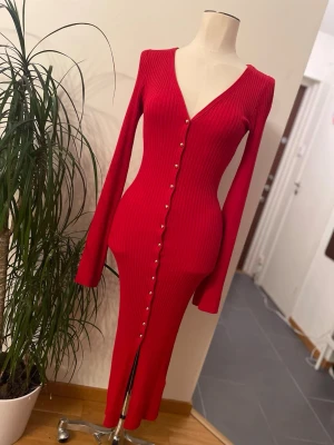 Röd ribbad långklänning med knappar - Säljer en snygg röd långklänning med ribbad struktur och v-ringad hals. Klänningen har långa ärmar och dekorativa knappar längs hela framsidan, samt en slits nedtill. Perfekt för dig som gillar en tight och figurnära passform.