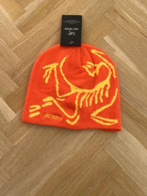 Orange mössa från Arc'teryx - Säljer en snygg orange mössa från Arc'teryx med ett stort gult logomotiv på framsidan. Mössan är mjuk och stretchig, perfekt för kalla dagar. Materialet är troligtvis syntet eller ullblandning. Passar dig som gillar färgstarka accessoarer.