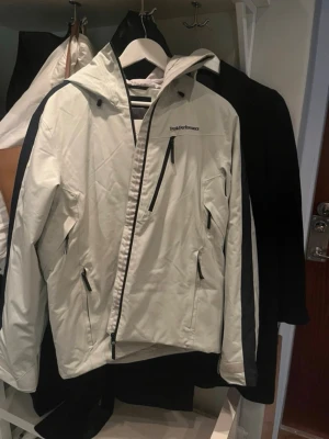 Vit vindjacka Peak Performance M - Snygg vit vindjacka från Peak Performance i storlek M. Jackan har huva, svarta detaljer och dragkedjor samt en cool svart logga på bröstet. Materialet är slitstark polyester och jackan har flera praktiska fickor med dragkedja. Perfekt för blåsiga dagar och outdoor.