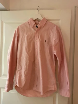 Rosa skjorta från Polo Ralph Lauren - Säljer en stilren rosa skjorta från Polo Ralph Lauren i slim fit. Skjortan har klassisk krage, knappar framtill och det ikoniska broderade logot på bröstet. Tillverkad i mjuk bomull och har långa ärmar. Perfekt för dig som gillar snygga och tidlösa plagg.