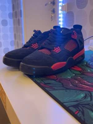 Nike Air Jordan 4 svart/röd sneakers - Säljer ett par Nike Air Jordan 4 sneakers i svart och rött med klassisk Jumpman-logga på hälen. Skorna har snörning, meshpaneler och detaljer i rött på sidorna och sulan. Materialet är mocka och syntet, med platt sula och rund tå. Perfekt för dig som gillar streetwear och ikoniska modeller.