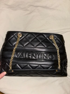 Svart quiltad axelväska från Valentino - Säljer en svart axelväska från Valentino med quiltat mönster och guldfärgade kedjor. Väskan har ett stort fack med dragkedja och flera mindre fack inuti. Materialet är skinn och den har en stilren, rektangulär form med tydlig logga framtill.