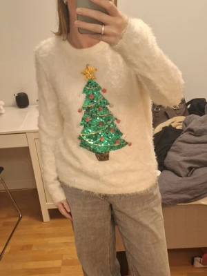 Fluffig jultröja med paljettgran - Mysig fluffig vit jultröja från My Favourite Xmas Jumper i storlek M. Tröjan har en stor paljettbroderad julgran med stjärna och kulor i grönt, guld, rött och silver på framsidan. Långärmad och riktigt mjuk, perfekt för julmys!