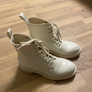 Vita boots med snörning i skinnimitation - Säljer ett par snygga vita boots med snörning och grov sula. Skorna är tillverkade i skinnimitation och har rund tå samt praktiska dragflikar bak. Perfekta för dig som gillar stilrena och robusta skor med lite edge.