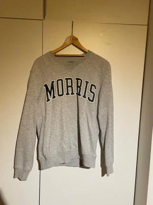 Grå sweatshirt från Morris, S - Säljer en grå sweatshirt från Morris i storlek S. Tröjan har klassisk rund hals, ribbade muddar och stor svartvit logotyp på bröstet. 