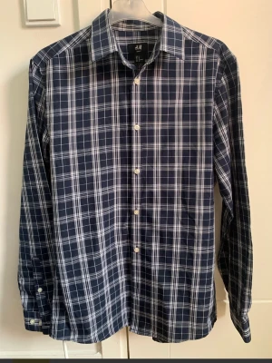 Blårutig slim fit skjorta från H&M stl M - Säljer en blårutig skjorta från H&M i slim fit-modell. Skjortan har klassisk krage, långa ärmar och vita knappar framtill. Materialet är en mix av 65% polyester och 35% bomull vilket gör den både mjuk och lätt. Perfekt för dig som gillar stilrena rutor.