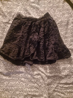 Svart volangkjol med struktur - Säljer en svart kortkjol med volangkant och snyggt strukturerat tyg. Kjolen har en elastisk midja som gör den bekväm och lätt att bära. Perfekt för dig som gillar en cool och avslappnad stil med lite extra detaljer.