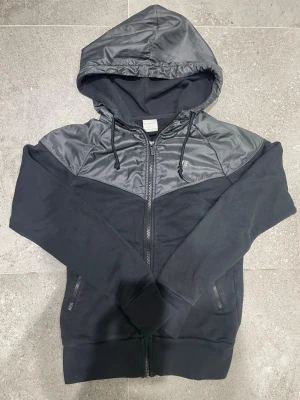 Svart vindjacka med huva från Nike - Nike vindjacka i svart med huva och dragkedja framtill. Jackan har snygga detaljer i blankt syntetmaterial över axlar och huva, samt snörning vid huvan. Två fickor med dragkedja och klassisk Nike-logga på bröstet. Skicket är på topp absolut inga fel eller liknande! Skulle säga att den är lite liten för sin storlek så den funkar också för XS.  Pris går att diskutera☺️