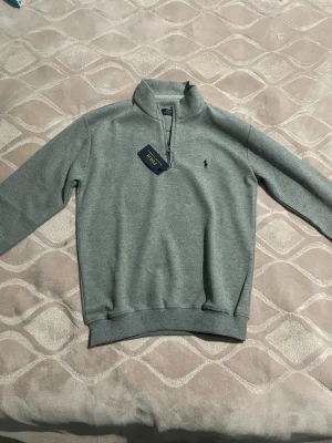 Grå half zip tröja från Polo Ralph Lauren - Snygg grå långärmad tröja från Polo Ralph Lauren med half zip och klassisk krage. Tröjan har en liten broderad logga på bröstet och är gjord i mjuk bomull. Perfekt för dig som gillar stilrena och bekväma plagg.