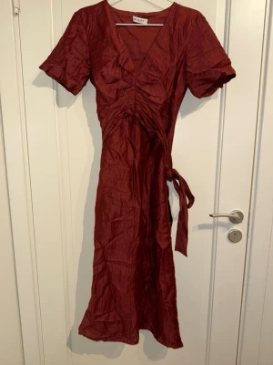 Vinröd omlottklänning från Masscob - Snygg vinröd långklänning från Masscob med omlottdetalj och knyt i midjan. Klänningen har korta ärmar, v-ringad hals och är i linne.