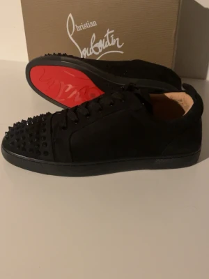 Svarta Christian Louboutin sneakers med nitar - Säljer ett par svarta sneakers från Christian Louboutin med ikonisk röd sula och svarta nitar på tån. Skorna har snörning och är tillverkade i skinn med en platt sula. Grymt snygga och sticker ut med sin edgy design och exklusiva känsla.