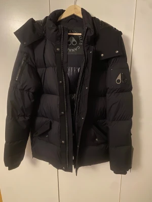 Svart pufferjacka från Moose Knuckles - Säljer en svart pufferjacka från Moose Knuckles för man i storlek XS med ikonisk logga på ärmen och snygga detaljer som dragkedjor och tryckknappar. Jackan har huva, långa ärmar och quiltad design. Den är endast använd 2-3 gr. Inköpt på thernlunds för 9000 :-, säljer den för 4500 :- (priset kan diskuteras vid snabbköp) 