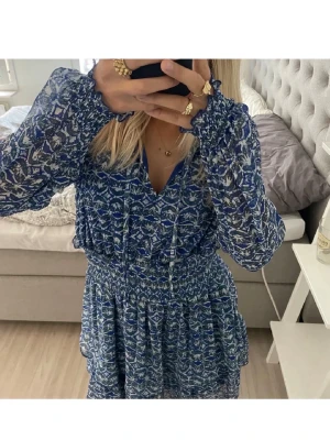 Blå mönstrad kortklänning från Zara - Säljer en blå och vit kortklänning från Zara med långärmade transparenta ärmar och ett snyggt grafiskt mönster. Klänningen har v-ringning, smock i midjan och volangdetaljer. Inga defekter alls! 