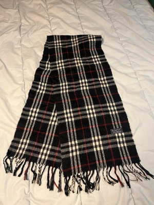 Rutig svart halsduk från Burberrys - Snygg rutig halsduk från Burberrys i svart, vit och röd. Västen är tillverkad i 100% lammull och har klassiska fransar längst ner. Den har ett stilrent Burberrys-märke och är perfekt för dig som gillar tidlös design med lite edge. N33