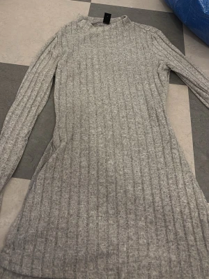 Grå ribbad stickad tröja - Säljer en grå långärmad tröja med ribbad struktur och omlott-detalj nedtill. Tröjan är stickad och har en hög hals som ger en snygg och modern look. Perfekt för dig som gillar stilrena och bekväma plagg.