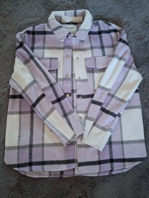 Rutig overshirt från ONLY i lila - Säljer en snygg rutig overshirt från ONLY i lila, vitt och grått. Jackan har klassisk krage, tryckknappar framtill och två bröstfickor. Materialet är mjukt och känns lite ulligt, perfekt för lager-på-lager. Passar dig som gillar chill och trendig stil.