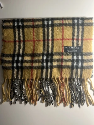 Rutig beige Burberry väst i ull - Säljer en klassisk rutig väst från Burberry i beige, svart, vitt och rött. Västen är tillverkad i 100% lammull och har fransar nedtill. Den har det ikoniska Burberry-mönstret och är mjuk och varm, perfekt för lager-på-lager-stil. (9B)