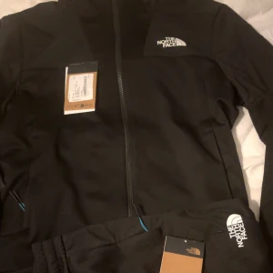 Svart vindjacka från The North Face - Svart vindjacka från The North Face med dragkedja framtill och logga på bröstet. Jackan har långärmade ärmar och Mountain Athletics-tryck på ena armen. Materialet är polyester och har en sportig, strukturerad yta. Perfekt för aktiva dagar utomhus.  STORLEK M OCH L FINNS MÖTS UPP STOCKHOLM