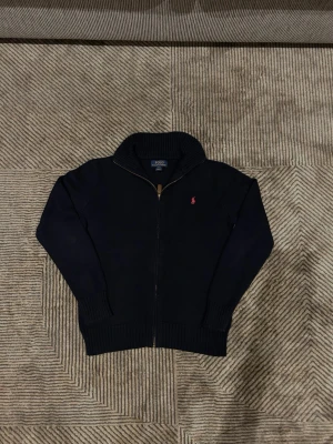 Ralph lauren stickad helzip - Säljer en marinblå stickad kofta från Polo Ralph Lauren med dragkedja och ribbad krage. Klassisk röd logga broderad på bröstet och långa ärmar. Perfekt till höst och vinter för en stilren look.