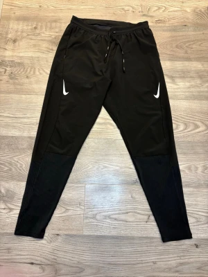 Svarta Nike träningsbyxor med snörning - Säljer ett par svarta träningsbyxor från Nike med vita loggor på båda benen. Byxorna har snörning i midjan och ribbade detaljer nedtill på benen. Tillverkade i ett lätt och stretchigt syntetmaterial som passar perfekt för träning eller chill.