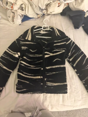 Svart teddyjacka med zebramönster H&M XS - Säljer en svart teddyjacka från H&M i storlek XS med fett zebramönster i vitt. Jackan har dragkedja framtill och är gjord i mjukt fleece-material. Perfekt för dig som gillar att sticka ut med coola mönster och vill ha något riktigt mysigt.