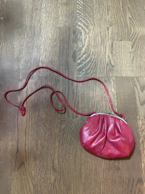 Röd kuvertväska i skinn - Söt röd kuvertväska i skinn med lång axelrem och metallspännet. Väskan har en mjuk, rundad form och rynkade detaljer framtill. Insidan är fodrad i matchande rött tyg. Perfekt för att bära det viktigaste och ger en retrovibe till din outfit.