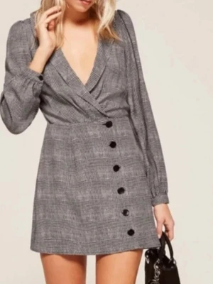 Rutig kortklänning från Reformation - Snygg kortklänning från Reformation i svart och grått rutigt mönster. Klänningen har omlott-design med djup v-ringning, långa ärmar och knappar framtill. Den har även ett knytband i midjan för extra passform. Perfekt för dig som gillar trendiga och klassiska plagg.