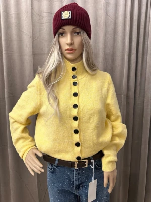 Gul stickad kofta med svarta knappar - Superfin gul stickad kofta med höga muddar och svarta knappar hela vägen framtill. Koftan har en hög krage och är långärmad, perfekt att slänga på sig för en färgklick i vardagen. Mjuk och skön känsla, passar till jeans eller kjol.