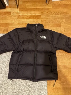 North face puffer jacka nuptse 1996 - Säljer en svart pufferjacka från The North Face med broderad logga på bröstet. Jackan har hög krage, dragkedja framtill och två sidofickor. Den är quiltad och har justerbara snören vid nederkanten. Perfekt för kalla dagar.