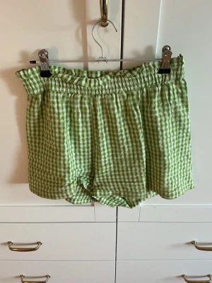 Gröna rutiga shorts med resårmidja - Jag säljer ett par gröna och vita rutiga shorts med högmidjad resår och volangkant upptill. De är gjorda i ett lätt och luftigt bomullstyg, perfekta för varma dagar. Passformen är loose och de har en avslappnad vibe. Köparen står för frakten.