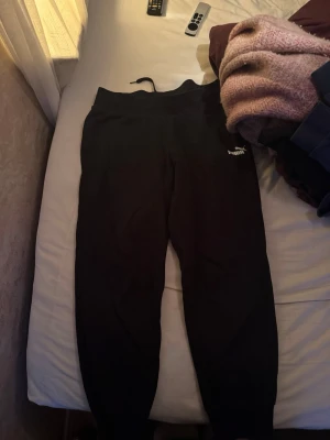 Svarta mjukisbyxor från Puma, M - Svarta mjukisbyxor från Puma i storlek M. Klassisk design med ribbade muddar vid benslut och elastisk midja. Puma-logga i vitt på ena benet. Perfekta för chill eller träning, tillverkade i mjukt bomullsmaterial för extra komfort.