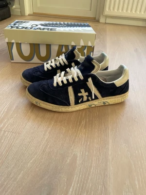 Premiata sneakers i mörkblå mocka - Säljer ett par Premiata sneakers i mörkblå mocka med vita detaljer och snörning. Skorna har en rund tå, platt sula med tryck och kontrasterande vita partier på häl och sidor. Snyggt broderad logga på sidan och coolt mönstrad sula. Box medföljer.