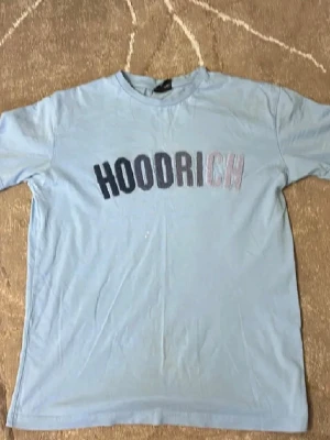 Ljusblå HOODRICH t-shirt - Säljer en ljusblå t-shirt från HOODRICH med tryckt logga i olika färger över bröstet. Klassisk rund hals och korta ärmar. Perfekt för en avslappnad streetwear-look.