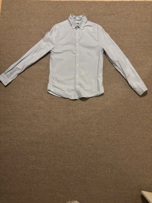 Ljusblå skjorta Dressmann  - Snygg ljusblå skjorta från Dressmann i slim fit-modell. Skjortan har klassisk krage, långa ärmar och knappar framtill. Perfekt för dig som gillar stilrena och enkla plagg. Materialet är mjuk bomull som känns skönt mot huden. Är använd få tal gånger 