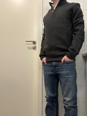 Mörkgrå stickad polotröja Pier One - Snygg mörkgrå stickad polotröja från Pier One med half zip och ribbad krage. Tröjan har lång ärm och är gjord i mjukt material som håller dig varm. Perfekt till jeans eller chinos, och dragkedjan har en stilren läderdetalj.