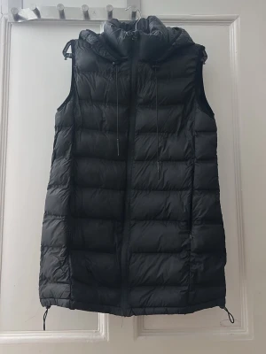 Svart lång dunväst med huva - Säljer en svart, lång dunväst med huva och dragsko både i midjan och vid huvan. Västen har quiltad design och dragkedja framtill. Perfekt för lager på lager och riktigt snygg till höst- och vinteroutfits.