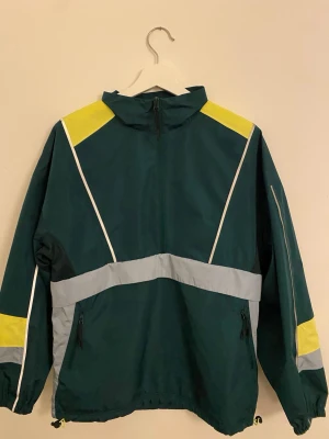 Sportig vindjacka med reflexdetaljer ASOS – stl S (oversize) - Säljer en snygg vindjacka från ASOS i mörkgrönt med gula och grå detaljer. Jackan är oanvänd i mycket bra skick, har halv dragkedja, hög krage och två fickor med dragkedja. Perfekt för dig som gillar sportig stil och vill sticka ut med färg. Justerbar nederkant med snörning.