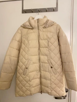 Beige quiltad pufferjacka – Henry Holly – stl S - Beige quiltad pufferjacka från Henry Holly med huva och dragkedja framtill. Jackan har två fickor med knappstängning och är varm utan att kännas klumpig. Använd men i fint skick. Stilren modell som passar perfekt till vardags under kyligare dagar.