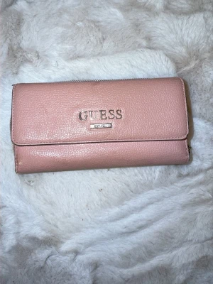 Rosa plånbok från Guess med dragkedja - Säljer en snygg rosa plånbok från Guess med silverfärgade detaljer och logga framtill. Plånboken har flera fack för kort, två fönster för ID och ett stort fack med dragkedja. Utsidan är i rosa skinnimitation och insidan är metallic-silver. Perfekt för dig som vill ha ordning på korten!