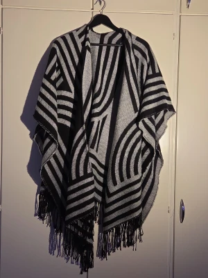 Svart och grå poncho - Säljer en snygg poncho i svart och grått med grafiskt randigt mönster. Ponchon har öppet framtill och långa fransar längs nederkanten. Perfekt att slänga över en outfit för extra stilpoäng. Materialet känns mjukt och stickat.