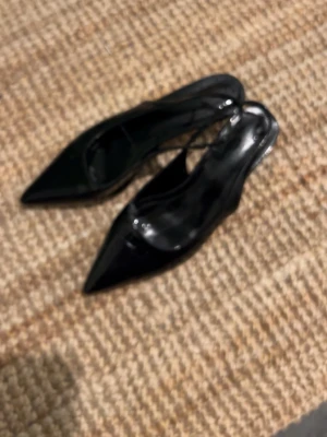 Svarta lackpumps med spetsig tå - Säljer ett par svarta pumps i glansigt lackmaterial med spetsig tå och låg klack. Skorna har en smal rem över hälen och en elegant, stilren design som passar till många olika outfits. Perfekt för dig som gillar klassiska och trendiga skor.