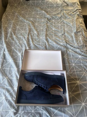 Mörkblå sneakers från Christian Louboutin - Säljer ett par mörkblå sneakers från Christian Louboutin med snygg röd sula och broderad logga på hälen. Skorna har snörning, rund tå och är tillverkade i mocka med beige innersula. Perfekt för dig som vill sticka ut med exklusiva detaljer.