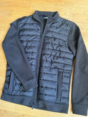 Svart quiltad pufferjacka Dressmann - Svart pufferjacka från Dressmann med quiltad framsida och släta ärmar. Jackan har dragkedja hela vägen, ståkrage och två sidofickor. Materialet är polyester och jackan har en clean, modern look som funkar till många stilar.