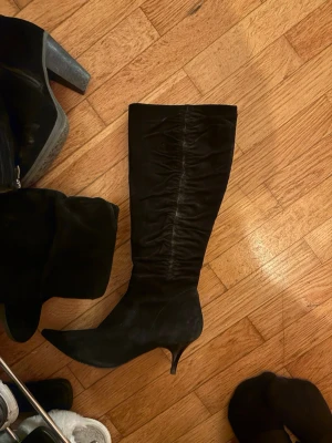 Svarta stövlar med spetsig tå och klack - Säljer ett par svarta höga stövlar i mocka med snygg rynkad effekt längs skaftet. Skorna har spetsig tå och smal klack som ger en elegant siluett. Perfekt för dig som vill ha en stilren och edgy look. Passar till både jeans och kjol.